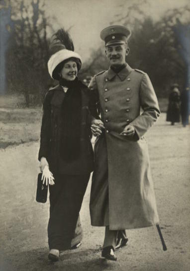 The prince and princess dressed in coats and hats, walking together in a park-like setting while holding hands and smiling.
