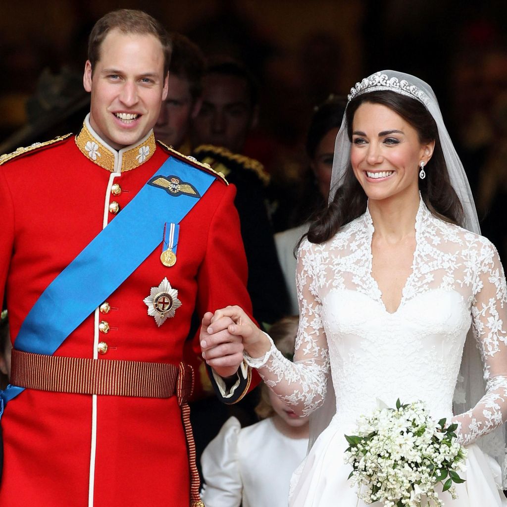 Photo of Prince William, dressed in red army tunic decorated with royal orders and Kate Middleton, dressed in white wedding gown and veil. In one hand she carries a bouquet of white flowers; her other hand is in Prince William's.