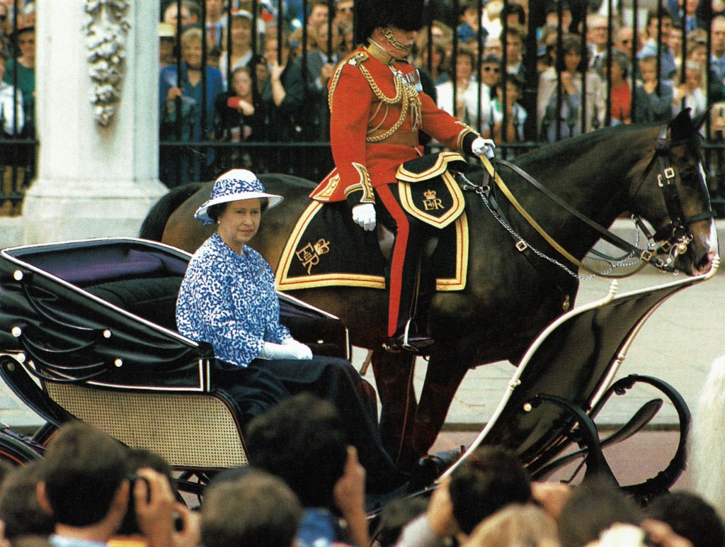 Photo of Queen Elizabeth riding in an open carriage. Beside her a uniformed member of the military rides on horseback.