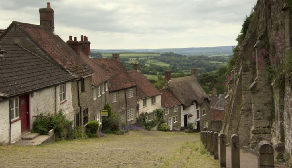Photo of a quaint village street with small cottages on the left and a stone wall on the right. In the background is green open landscape.