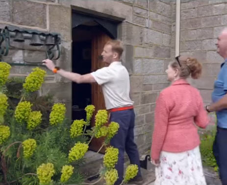 At the front door of a stone house, a man pulls the lever on a set of three bells mounted on the stone wall, while a man and woman watch.