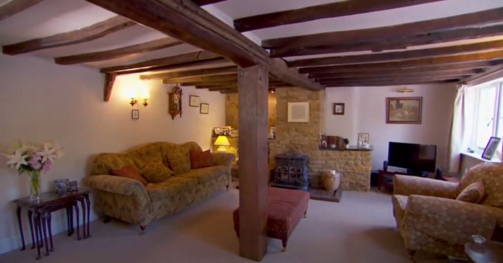Photo of a living or sitting room furnished with chair, sofa, tables, and television. In the center of the room is a large post that holds up a center beam that runs the length of the ceiling and has several perpendicular beams leading off of it.
