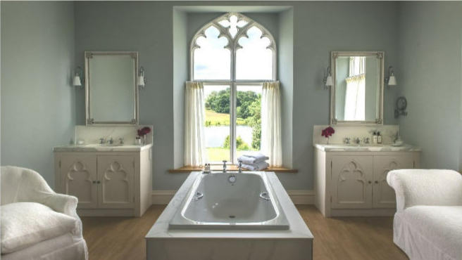 Photo of the interior of a large bathroom with a tall Gothic-framed window in the center of the exterior wall. A soaking tub is in the middle of the room. On either side of the window are twin vanities with sinks and mirrors. A white upholstered chair and a white chaise lounge stand on either side of the tub.