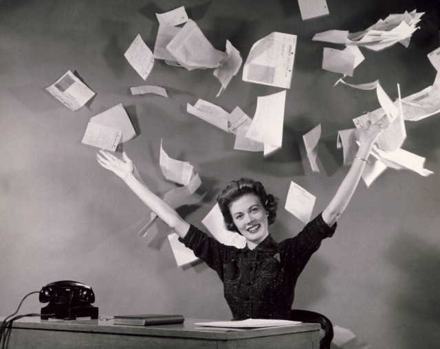 Black and white photo from 1960s of woman seated at desk on which is an old telephone, a book and some papers. She is smiling and holding her hands above her head, having just tossed a stack of papers up into the air. 