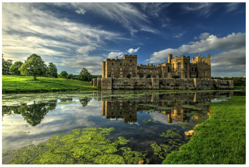 Long shot of medieval castle with long battlements across the front and protective walled walkways. The castle is set against lush parklands on one side and a tranquil lake on the other.